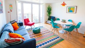 En lys stue med sofa, rød lænestol, turkise stole, et rundt bord og en farverig måtte.