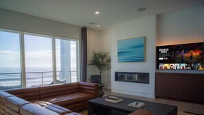 Ein modernes Wohnzimmer mit großem Fenster zum Meer, einem braunen Ledersofa, einem schwarzen Couchtisch und einem wandmontierten Flachbildfernseher.