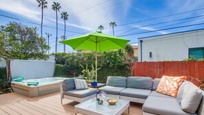 Eine Terrasse im Freien mit Whirlpool, einer grauen Couch, einem Korbstuhl-Tisch und einem grünen Sonnenschirm.