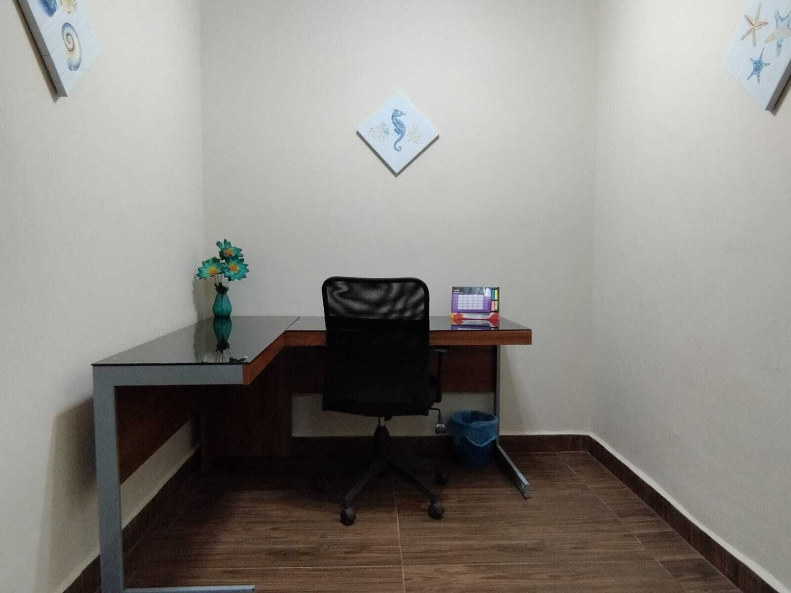 Un petit bureau comprenant un bureau, une chaise et un ordinateur. On trouve un vase avec des fleurs sur le bureau, et un tableau encadré au mur.
