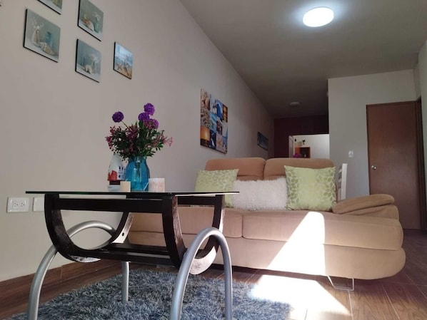A living room with a beige sofa, a coffee table, and a vase of flowers. There are wall-mounted pictures and a door in the background.