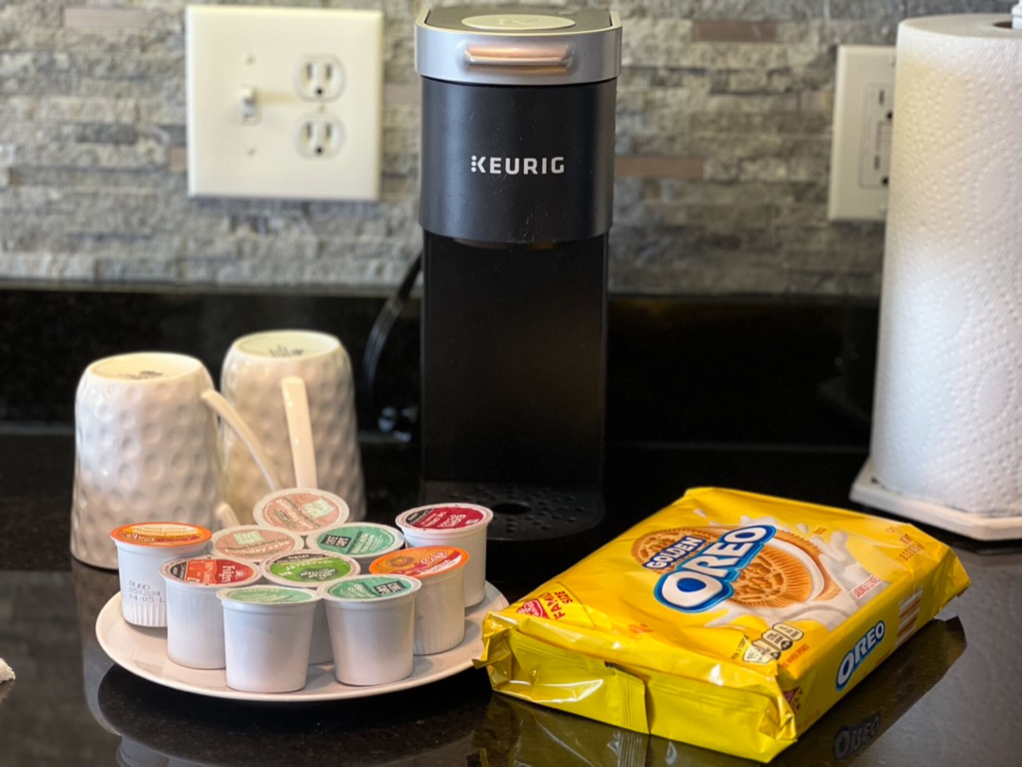 Uma bancada de cozinha com uma cafeteira Keurig, um prato com xícaras de café e uma embalagem de biscoitos Oreo.