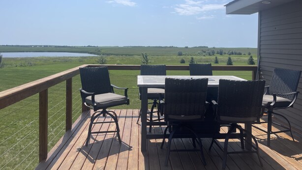 Een houten terras met een tafel en stoelen, uitkijkend op een grasveld en een waterpartij.