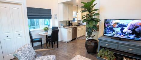A living room with a TV, a large potted plant, a patterned rug, and a dining area with a table and chairs.