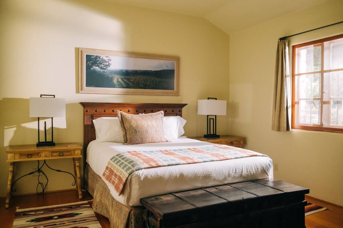 A bedroom with a bed, two bedside tables with lamps, a wooden bench, a rug, a window with curtains, and a framed landscape painting on the wall.
