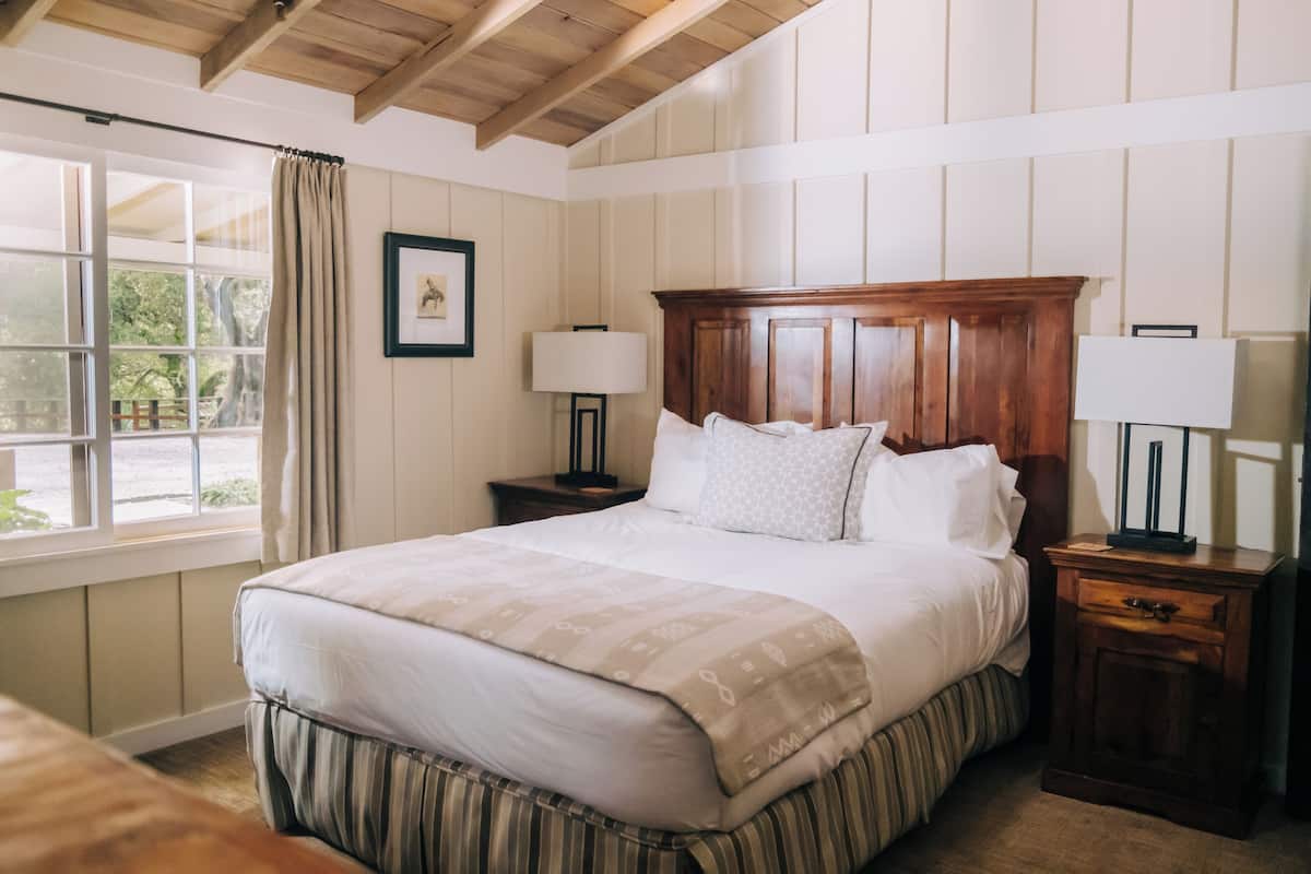A neatly made bed with a wooden headboard, two bedside tables with lamps, a window with curtains, and a framed picture on the wall.