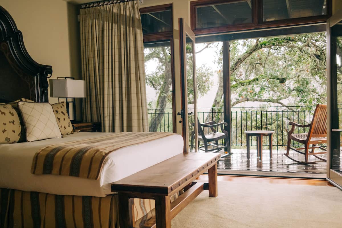 A bedroom with a bed, a bench, a nightstand, a lamp, and a view of the outdoors through a glass door.