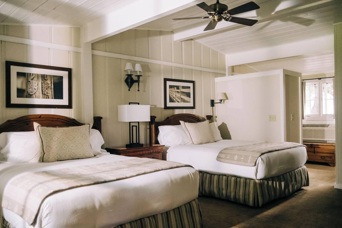 A hotel room with two beds, a ceiling fan, wall-mounted lamps, and framed pictures.