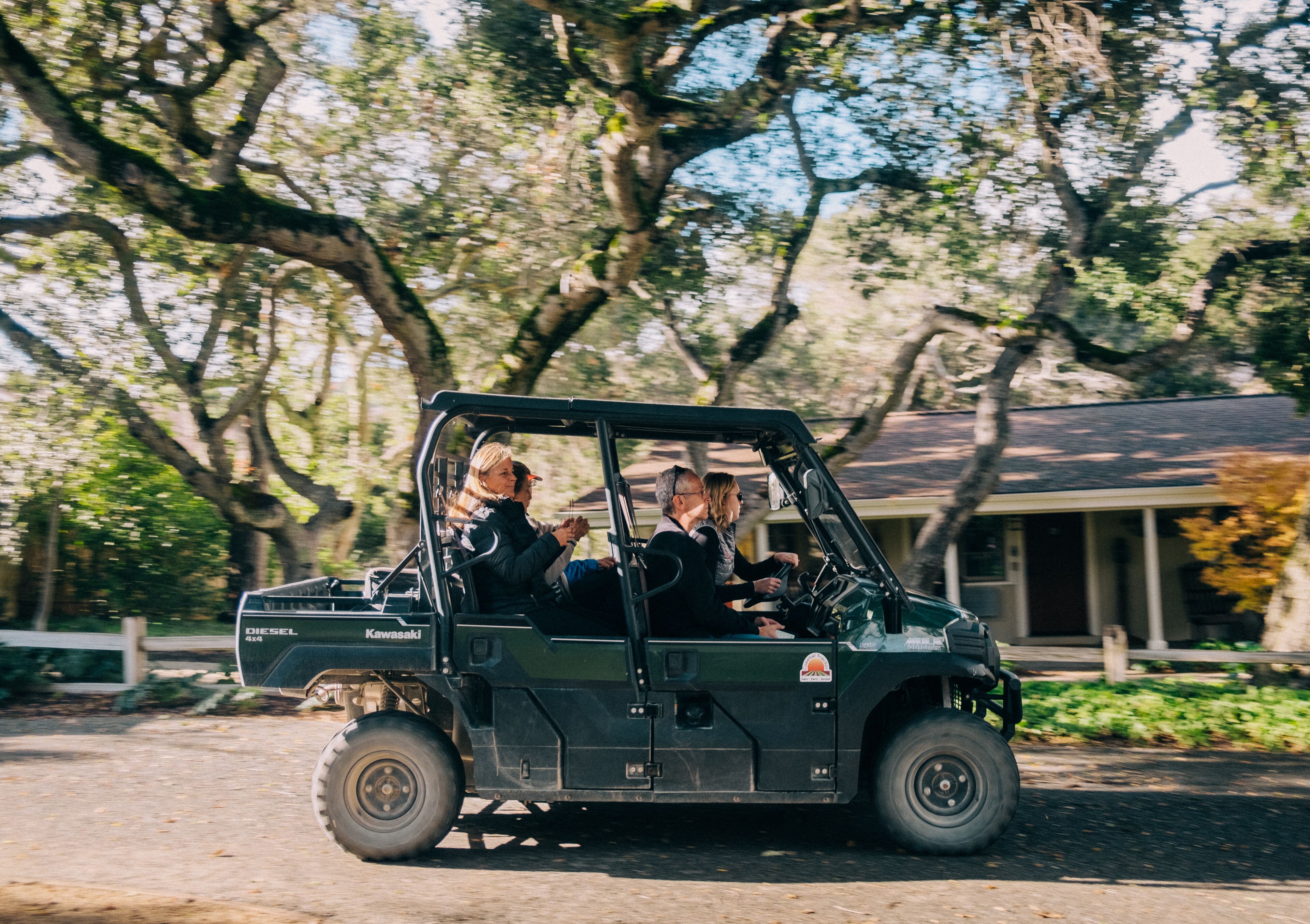 A green utility vehicle with two passengers, parked on a road with trees and a building in the background.