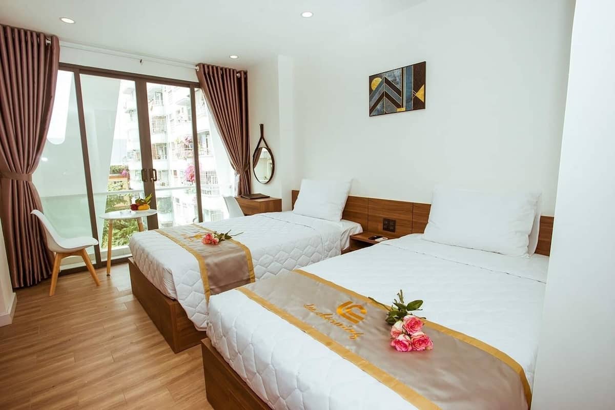A hotel room with two beds, a small table, a chair, and a view of the outside balcony.