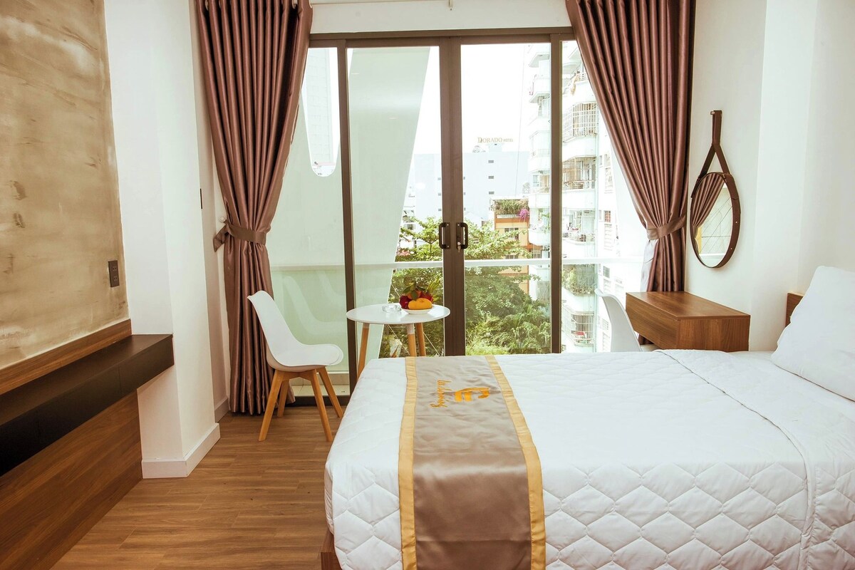 Ein Hotelzimmer mit Bett, einem kleinen runden Tisch mit Pflanze, einem Sessel und Blick auf die Stadt durch große Fenster.
