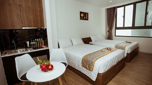 Ein Hotelzimmer mit zwei Betten, einer kleinen Küchenzeile, einer Essecke mit Tisch und Stühlen sowie einem großen Fenster mit Vorhängen.