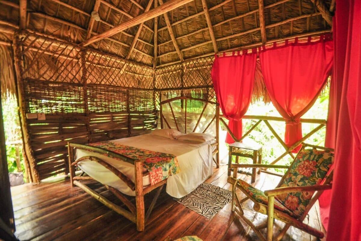 Una habitación con techo de paja, una cama, una silla y un ventanal con cortinas rojas.