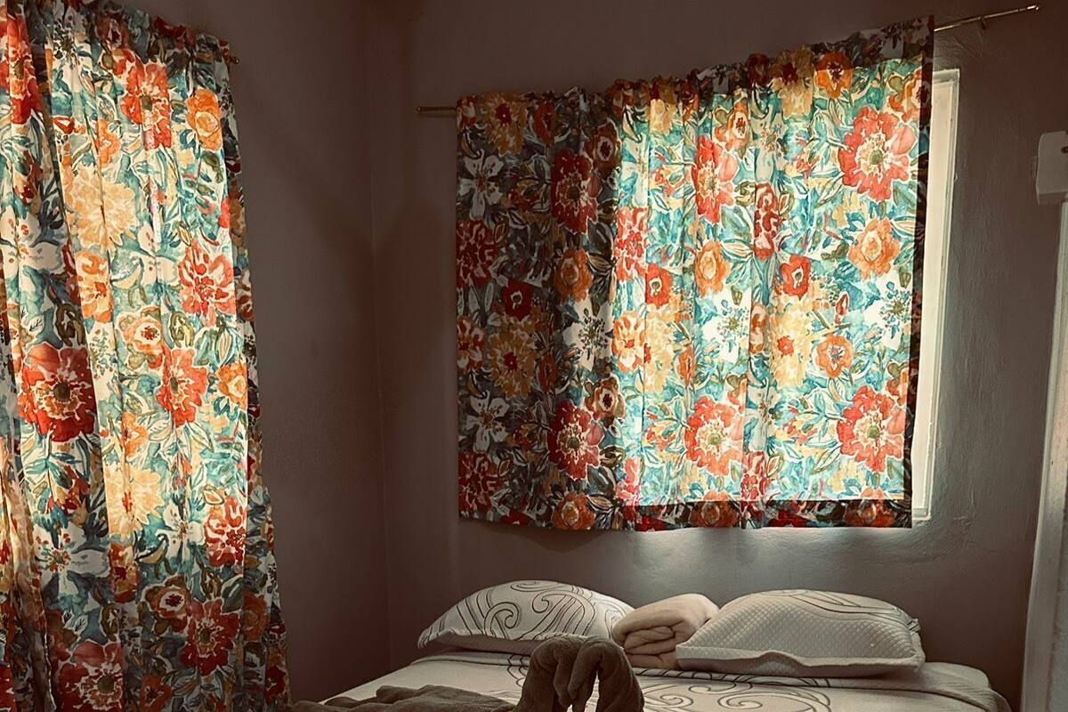 Una cama con cubrecama floral, una ventana con cortinas y un almohadón con diseño.