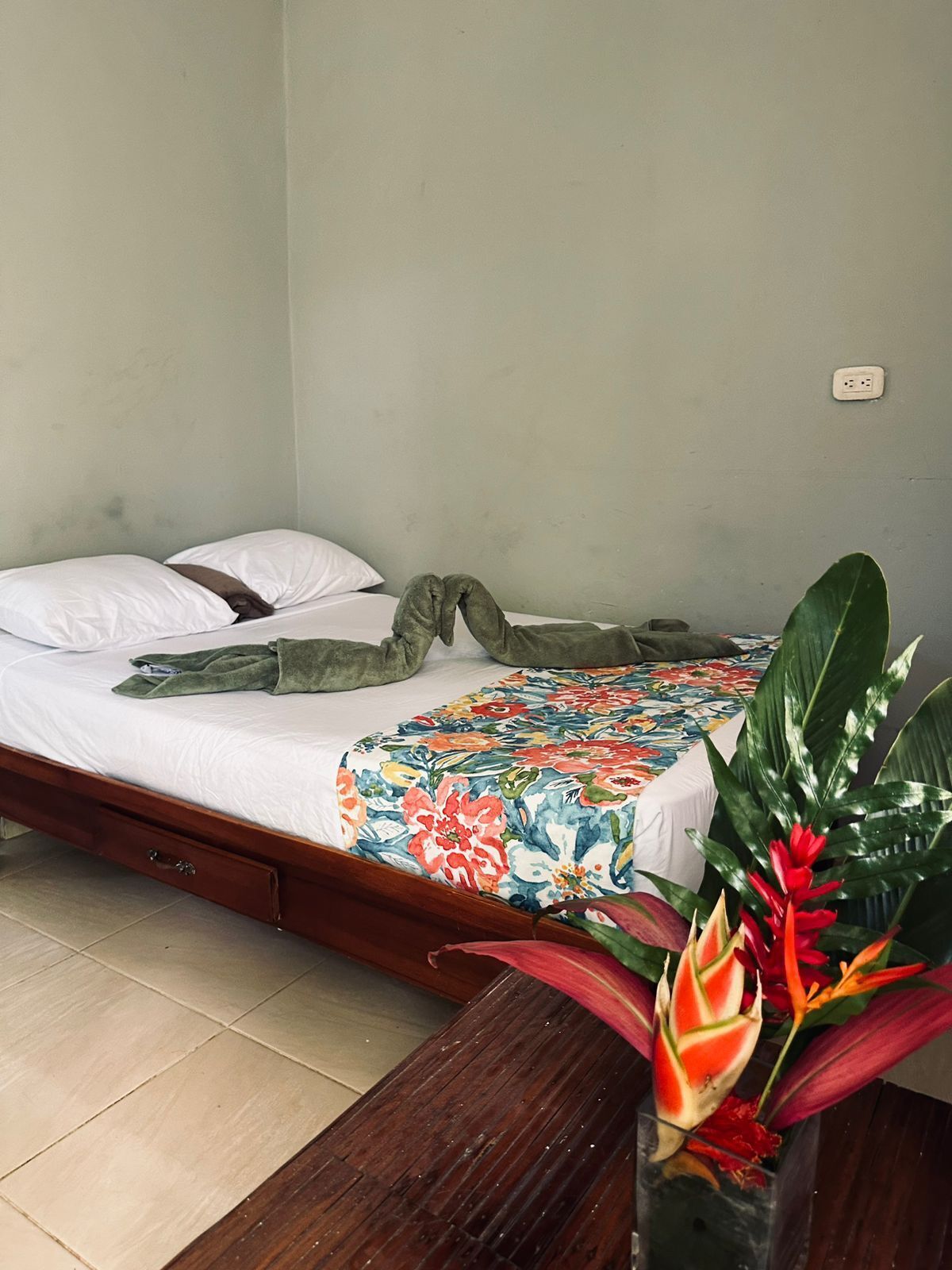Un lit simple recouvert d’une couvre-lit à motifs floraux, une serviette verte et une couverture verte. Il y a une table de chevet en bois avec une lampe et une prise électrique au mur. Une grande plante verte aux fleurs rouges se trouve dans un vase posé sur le sol.