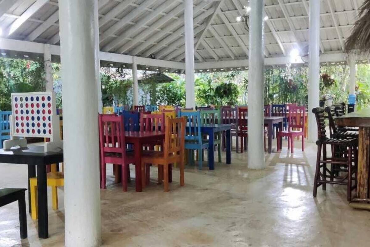 Un espacio al aire libre techado con sillas y mesas coloridas, una zona de bar y un techo de paja.