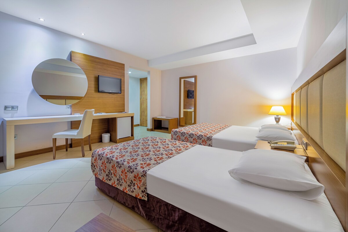 A hotel room with two beds, a desk, a chair, a TV, and a lamp.