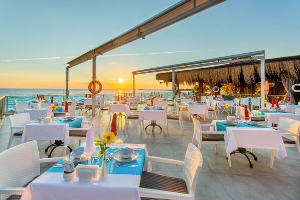 An outdoor dining area with tables set for two, overlooking the sea, featuring a thatched roof structure and a clear view of the sunset. - Justiniano Club Alanya (Alanya)