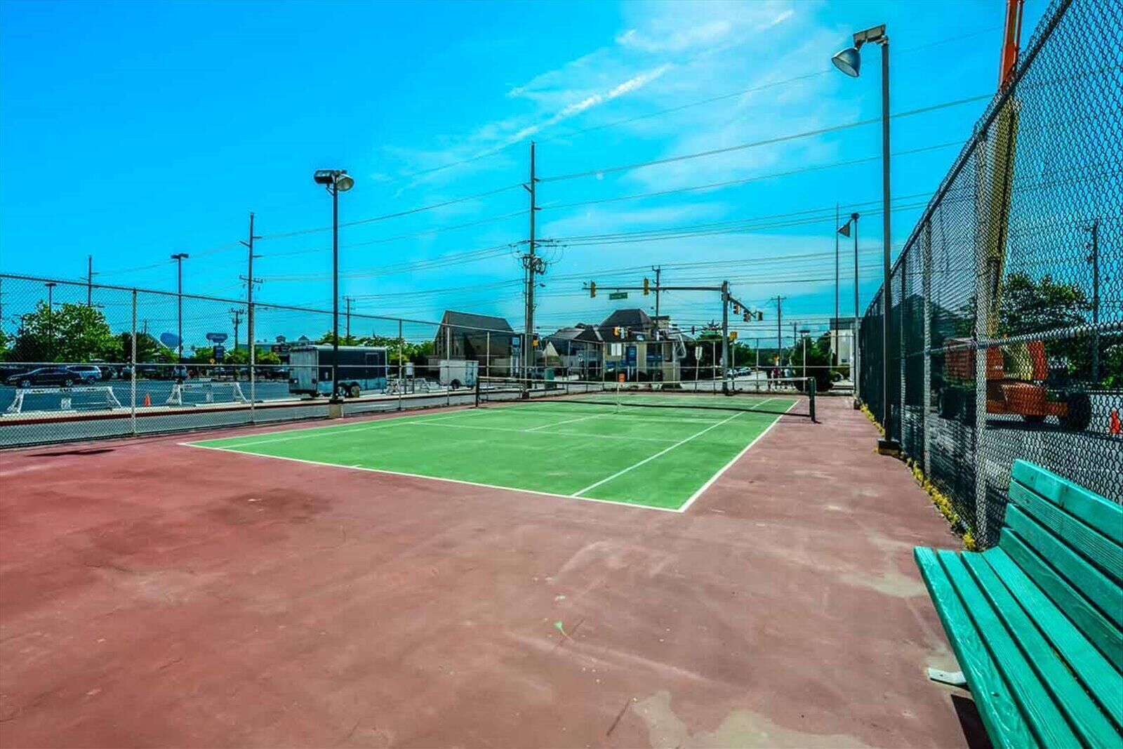 Ein Tennisplatz mit roter Belag, umgeben von einem Maschendrahtzaun, und eine grüne Bank auf der rechten Seite.