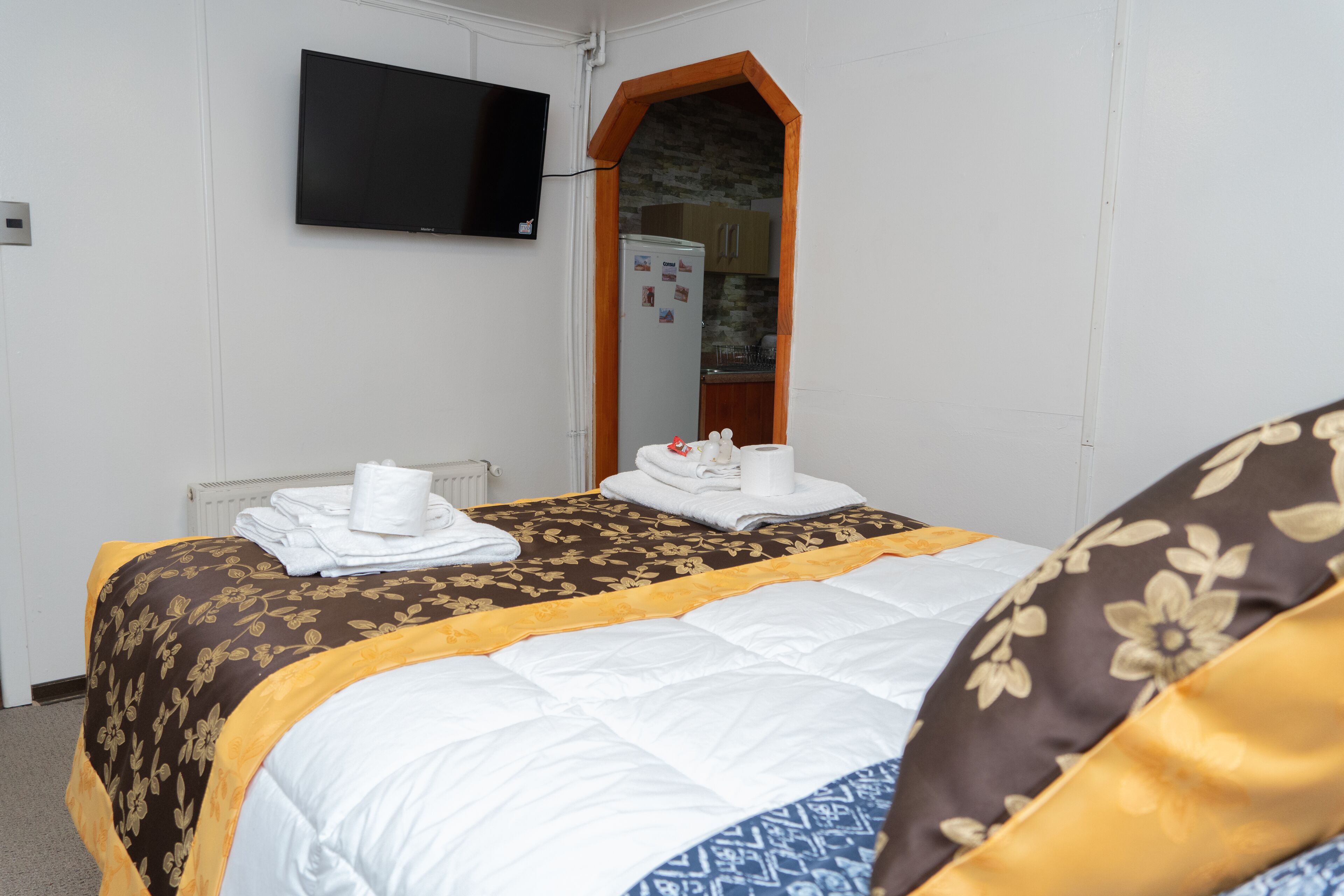 Habitación de hotel con cama, televisor montado en la pared, espejo y una cocina visible a través de un arco.