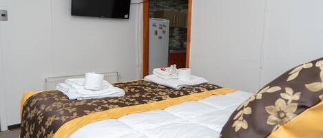 Habitación de hotel con cama, televisor montado en la pared, espejo y una cocina visible a través de un arco.