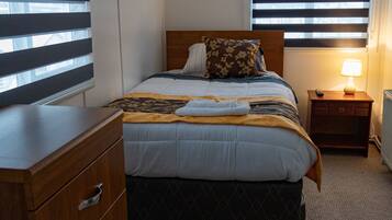 A hotel room with a bed, wooden dresser, bedside table with a lamp, and a window with blinds.
