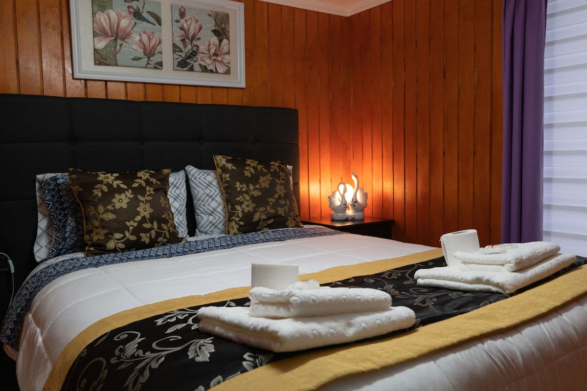 Ein Hotelzimmer mit einer holzverkleideten Wand, einem Bett mit blumigen Kissen, einem Nachttisch mit Kerze und Handtüchern auf dem Bett.