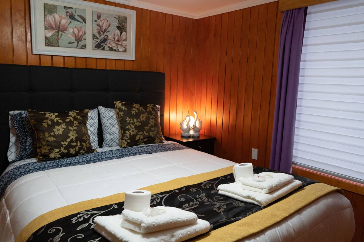 Ein ordentlich bezogenes Bett mit floralen Kissen, einer Nachttischlampe und einem Fenster mit Jalousien.