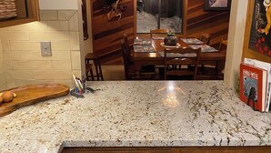 Una cocina con encimera de granito, armarios de madera, una mesa de comedor con sillas, una araña y un ventanal con vistas a la nieve.