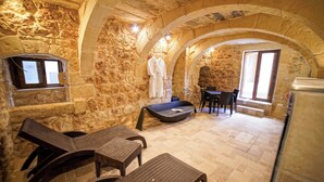 Une pièce aux murs de pierre, avec un banc de pierre, deux fauteuils, une table et une fenêtre.