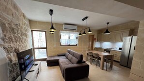 Een moderne woonkamer met een modulaire bank, een flatscreen televisie en een accentmuur van steen. Achtergrond toont een keuken met een eethoek en een koelkast.