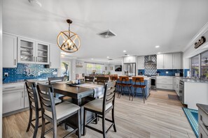 Cozinha moderna com armários brancos, uma ilha central com banquetas, um lustre e vista para o exterior.