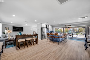 Uma sala de estar ampla com área de jantar de madeira, cozinha com uma ilha grande e vista para a área da piscina.