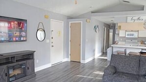 A living room with a grey sofa, a TV, and a wooden entertainment unit. There is a door leading to another room and a kitchen area with a microwave and cabinets. - Spacious Condo Near Disney (Kissimmee)