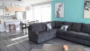 A modern living room with a grey sectional sofa, a coffee table, and a kitchen area in the background. - Spacious Condo Near Disney (Kissimmee)