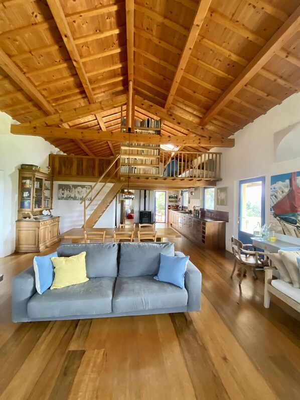 Uma sala de estar ampla com teto de madeira alto, uma estante embutida, um sofá com almofadas e uma escada que leva a um andar superior.
