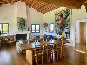 Uma sala de estar ampla com uma grande mesa de jantar de madeira, assentos confortáveis, lareira e um quadro abstrato vibrante.