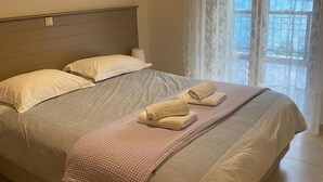 A neatly made bed with pillows and a folded towel, a headboard, and a window with curtains. - Secret Arkadia Bay Beachfront, Sea & mountain View, Secret Arkadia Bay Beachfront, Sea & mountain Vi (Arkadiko Chorio)