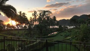 Campo de golf con vistas a la puesta de sol sobre un cuerpo de agua, rodeado de palmeras y vegetación.
