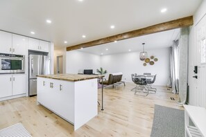 Cocina moderna con armarios blancos, electrodomésticos de acero inoxidable y una isla de madera. Cuenta con un comedor con mesa y sillas, y un ventanal con cortinas.
