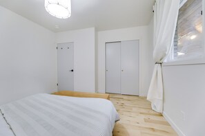 Un dormitorio con cama, suelo de madera, paredes blancas, dos puertas y una ventana con cortinas.