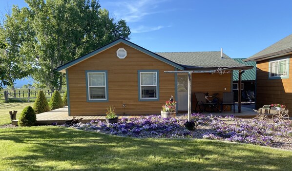 A small wooden house with two windows, a porch with outdoor seating, and a garden with purple flowers. - Adorable Out of Town Guest House (Hamilton)