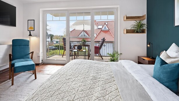 A bedroom with a large bed, a blue chair, a desk, and a balcony with a view of houses.
