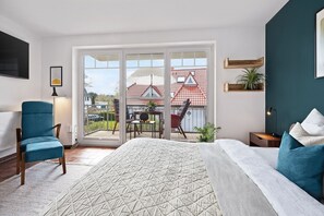 Une chambre avec un grand lit, une chaise bleue, un bureau et un balcon donnant sur des maisons.