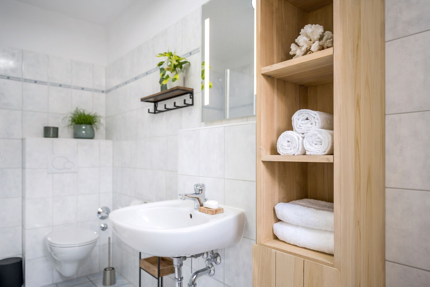 Une salle de bain moderne avec un lavabo blanc, des étagères en bois où sont posées des serviettes, des toilettes et une plante sur une étagère.