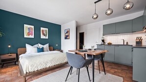 A modern bedroom with a bed, bedside tables, and a nightstand. There is a dining area with a table and chairs. The room has a kitchen area with cabinets and a countertop.