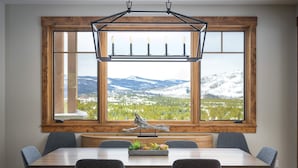 Un espace repas comprenant une grande table, six chaises, un lustre et une vue sur des montagnes enneigées, visible à travers une grande fenêtre.