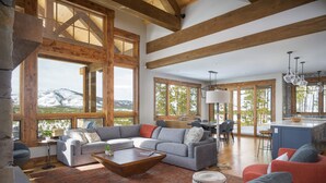 Un vaste espace de vie comprenant un grand canapé d’angle gris, un fauteuil rouge et une table basse en bois. De nombreuses fenêtres offrent une vue sur un paysage montagneux enneigé.