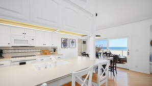 Une cuisine moderne avec des meubles blancs, un grand îlot central et une vue sur la plage grâce à une porte ouverte.