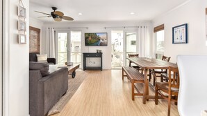 Ein großzügiges Wohnzimmer mit grauem Sofa, einem Holzespeisetisch und Stühlen. Ein Deckenventilator und ein Fernseher sind an der Wand angebracht.
